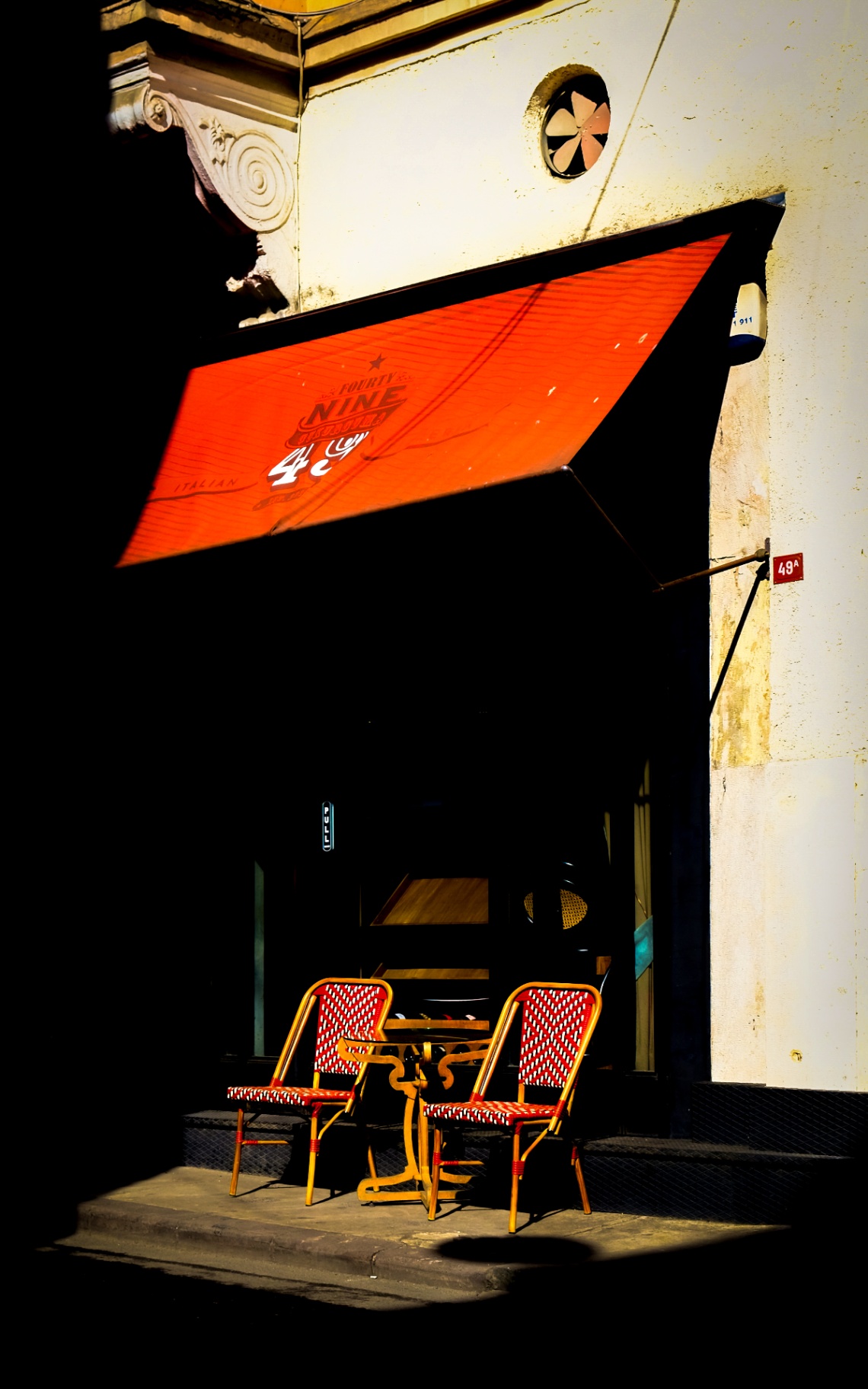Rode luifel boven twee stoelen in fel zonlicht voor een cafégevel.
