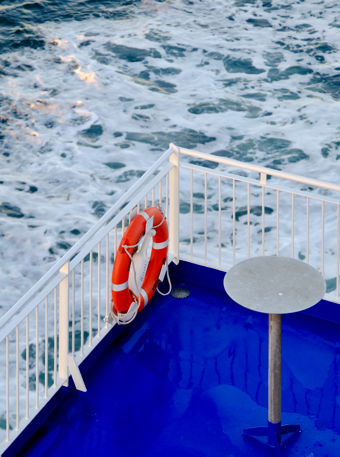 Oranje reddingsboei aan een witte reling op een blauw dek boven schuimend water.